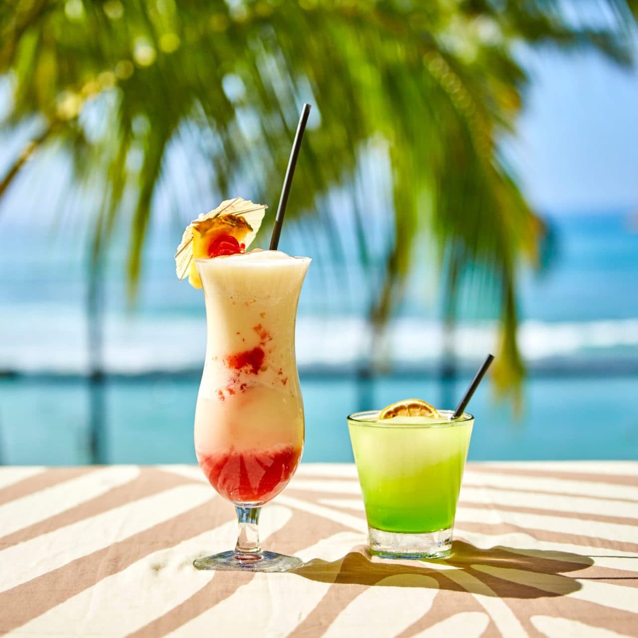 Tiki's Grill & Bar's classic Lava Flow cocktail with an umbrella and cherry, alongside a Melon Ball Cooler, on a table with a view of Waikiki Beach, waves, and palm trees.