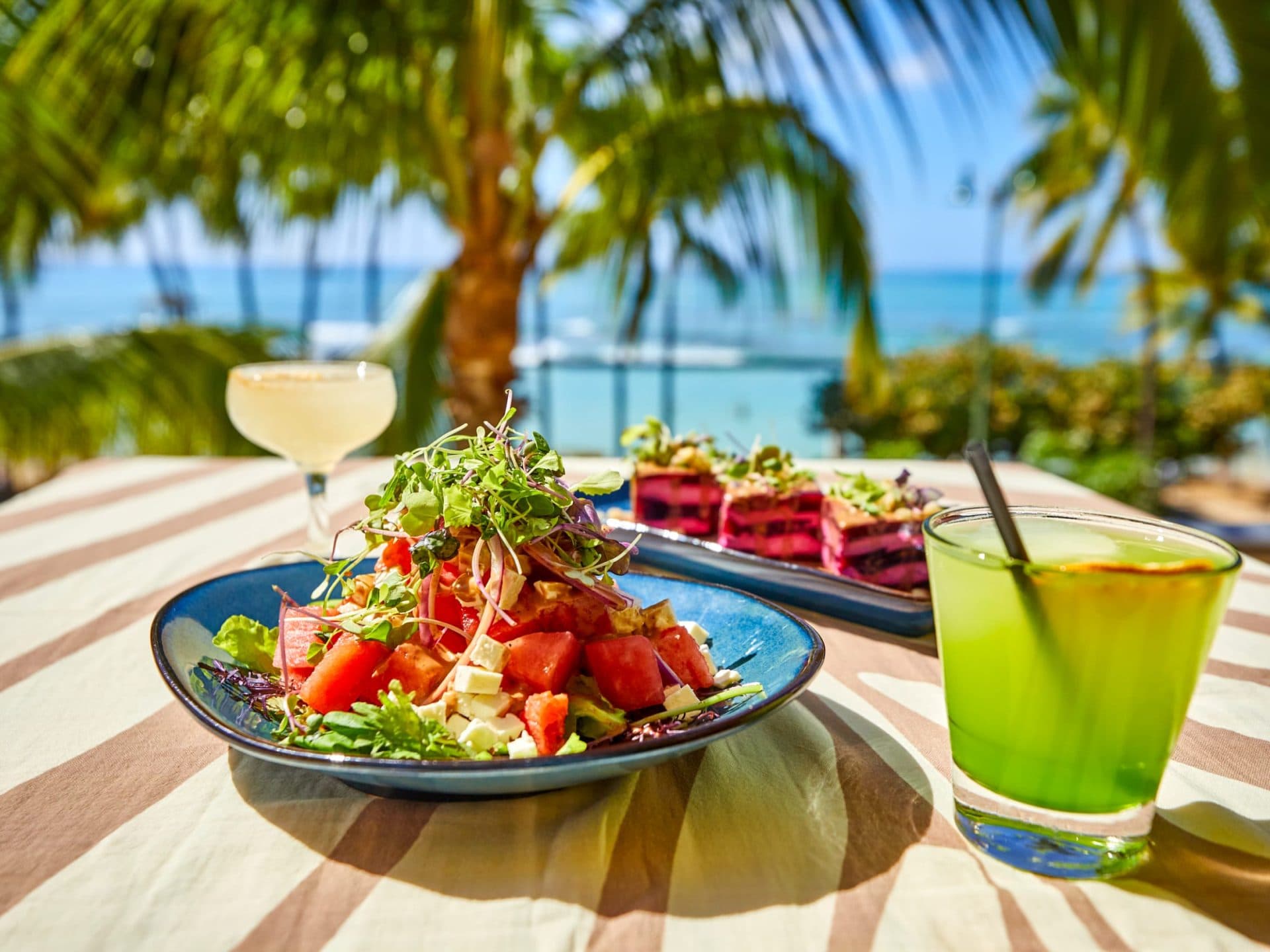 A fresh Farm-to-table Island Watermelon Salad on an outdoor table at Tiki's Grill & Bar, with an ocean view of Waikiki Beach, surf, and palm trees.