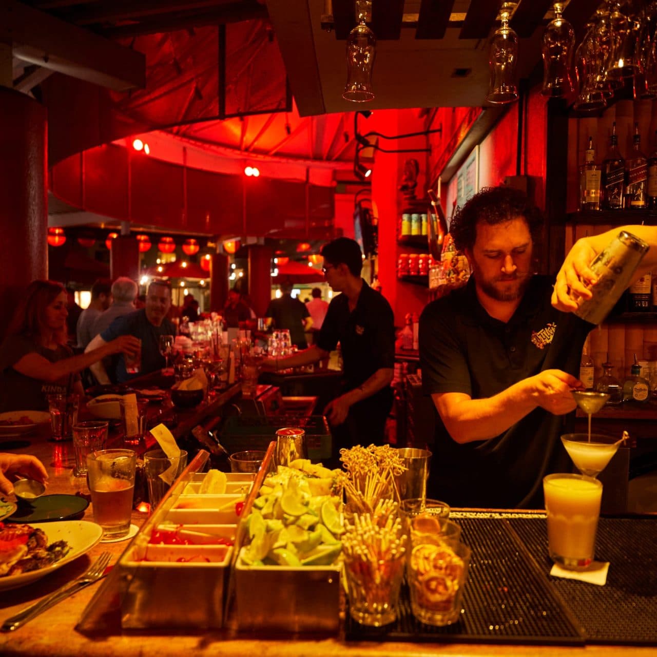 Bartender Austin at Tiki's Grill & Bar straining a craft cocktail, with a tray of garnishes and a lively nightlife scene in the background.