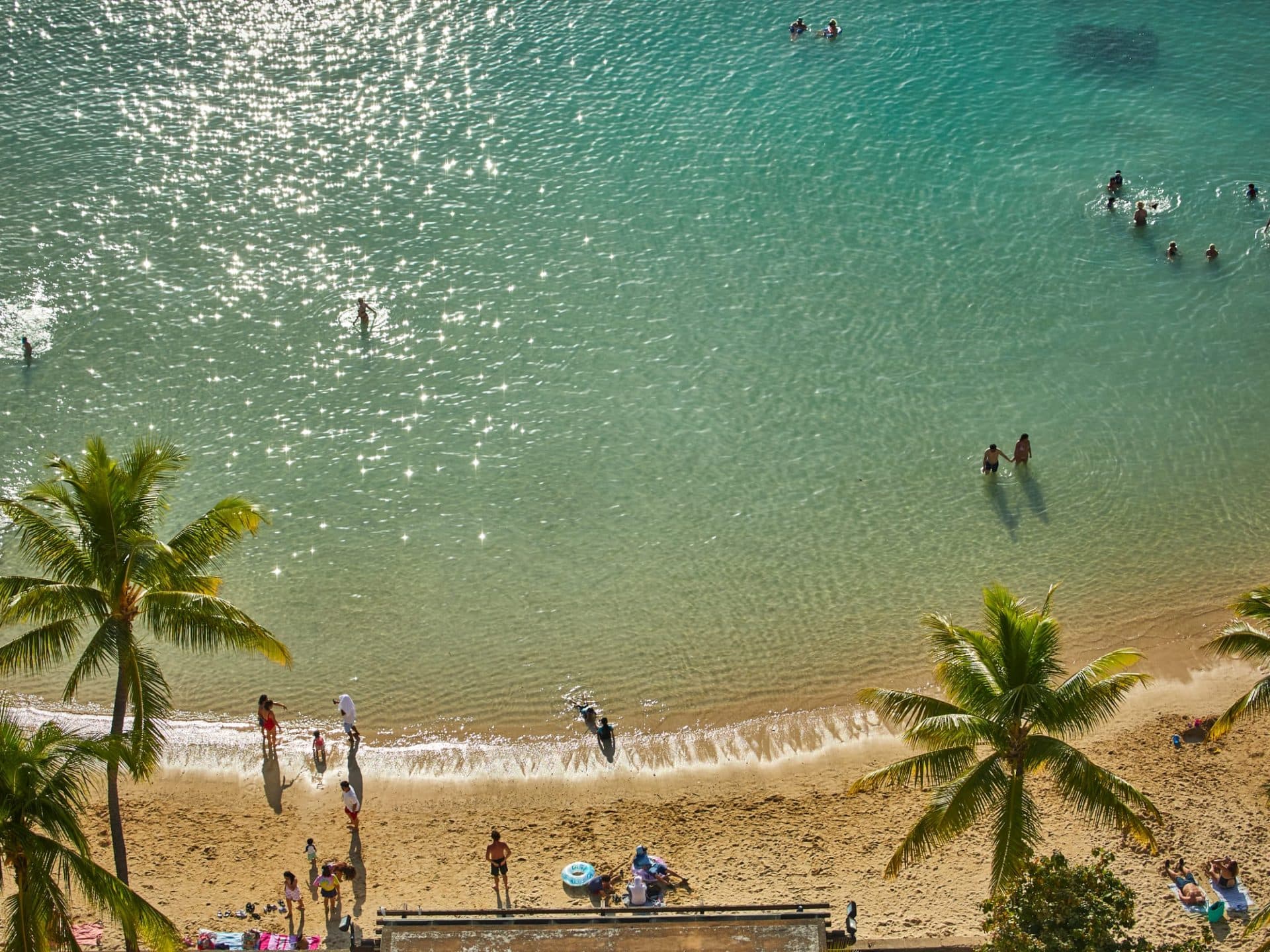A scenic photo of Waikiki Beach with families and friends enjoying the sand, with coconut and palm trees, showcasing a great people-watching spot.