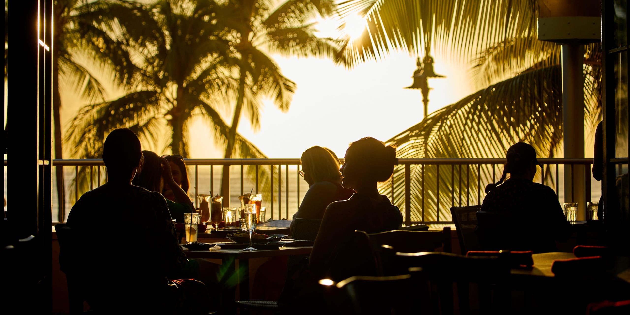 Silhouettes of guests at Tiki's Grill & Bar, enjoying tropical drinks and pupus with a beautiful sunset and ocean view in the background.