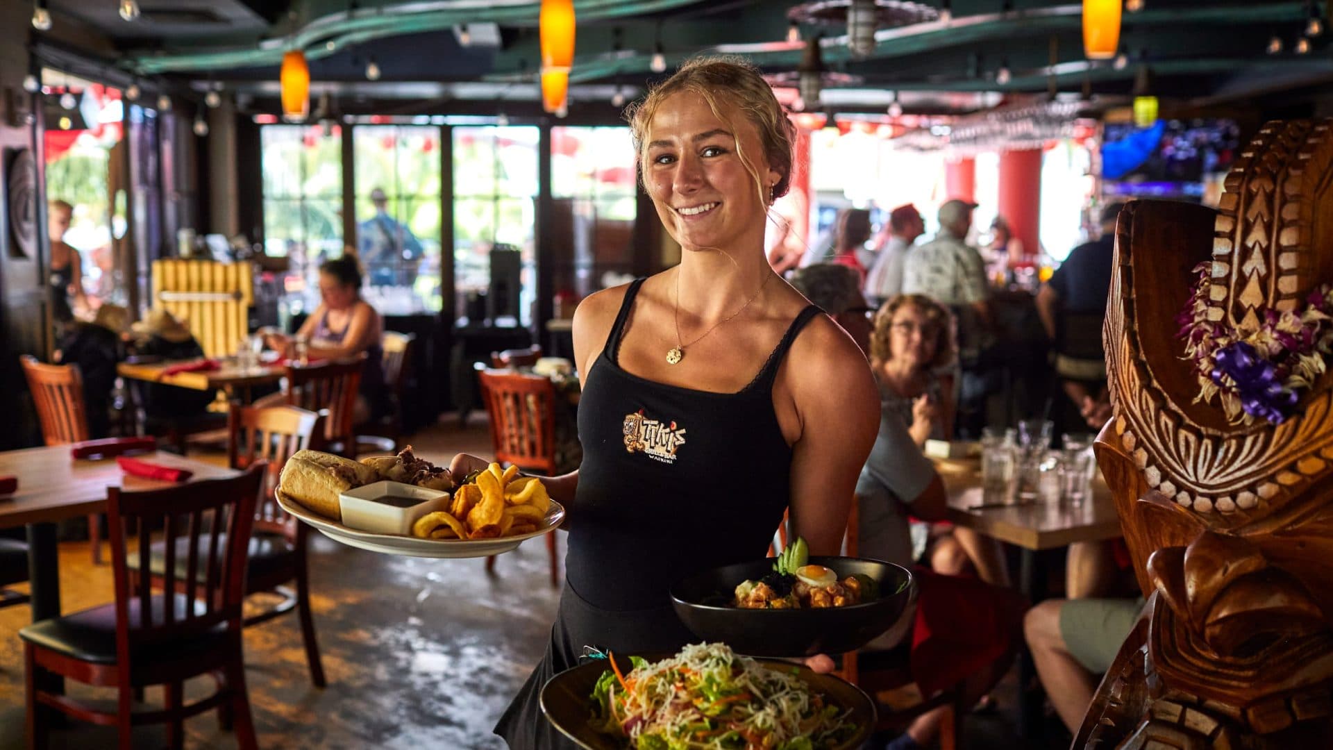 A smiling server with a tiki statue watching over her, serving a variety of dishes including Mama's Thai Chicken Salad, steak sandwich, and a poke bowl, showcasing the team's aloha spirit.