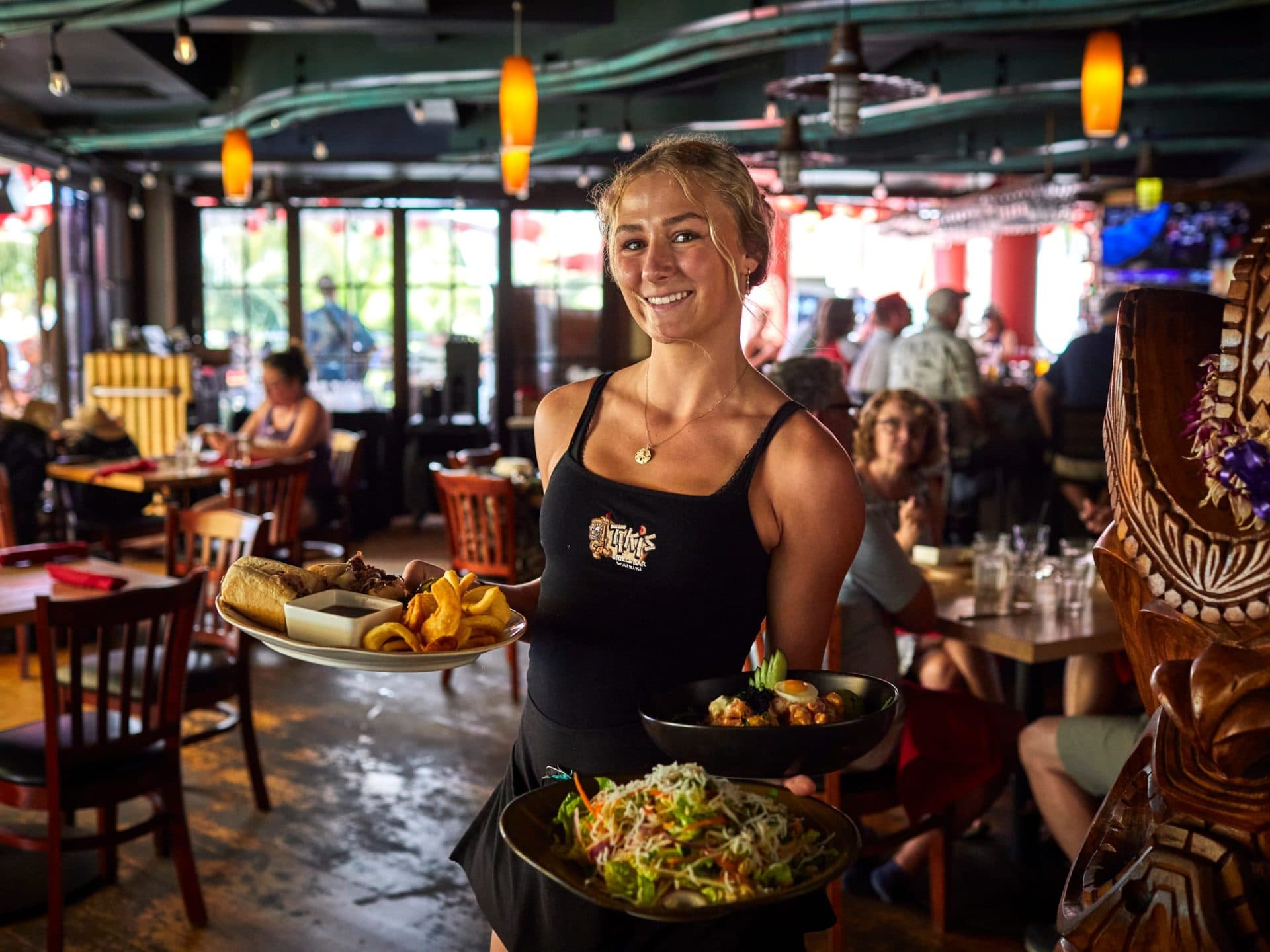 A smiling server with a tiki statue watching over her, serving a variety of dishes including Mama's Thai Chicken Salad, steak sandwich, and a poke bowl, showcasing the team's aloha spirit.