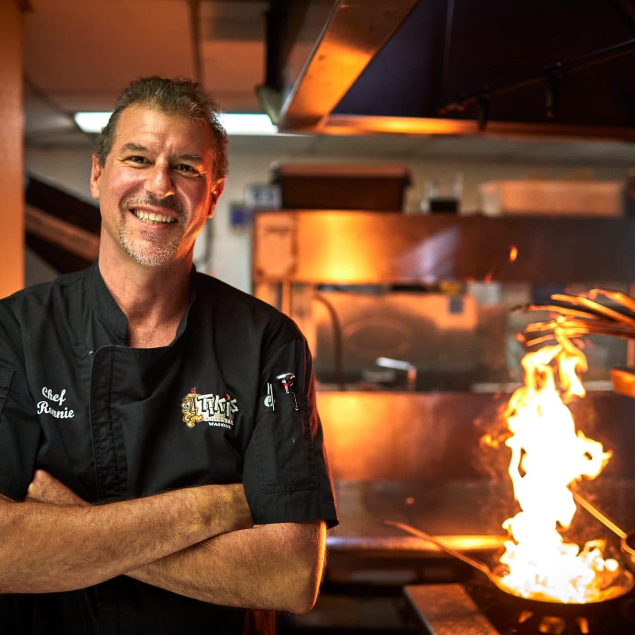 Executive Chef Ronnie Nasuti smiling in Tiki’s Grill & Bar kitchen with flames rising from the grill during a live cooking action shot.
