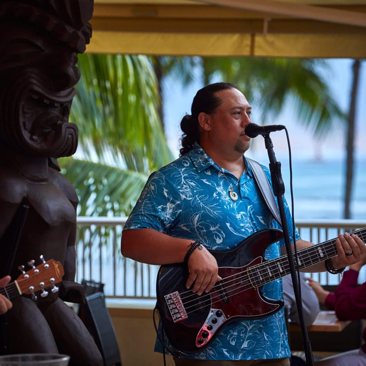 A local band playing live music at Tiki's Grill & Bar, with a big tiki statue, the beach, and the ocean visible in the background.