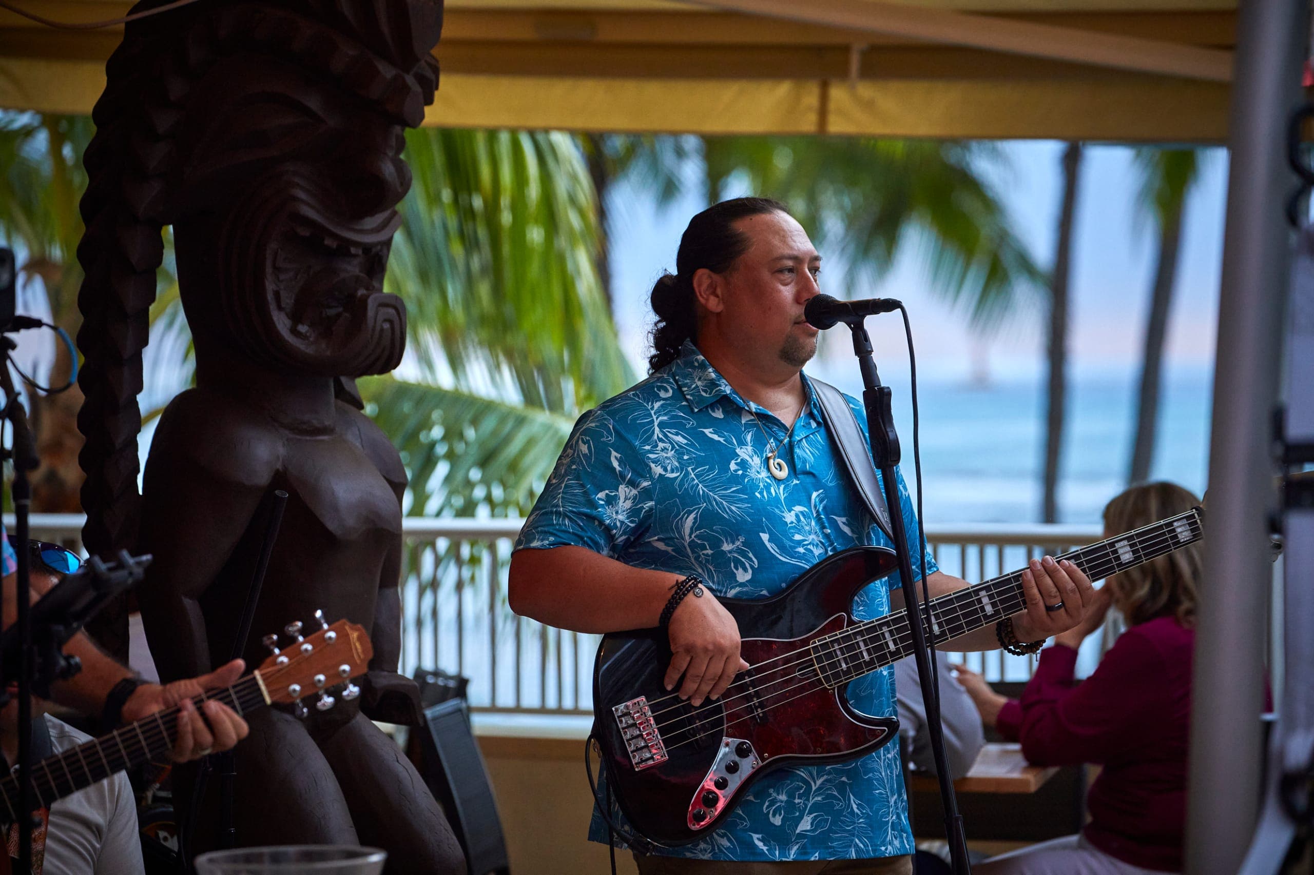 A local band playing live music at Tiki's Grill & Bar, with a big tiki statue, the beach, and the ocean visible in the background.