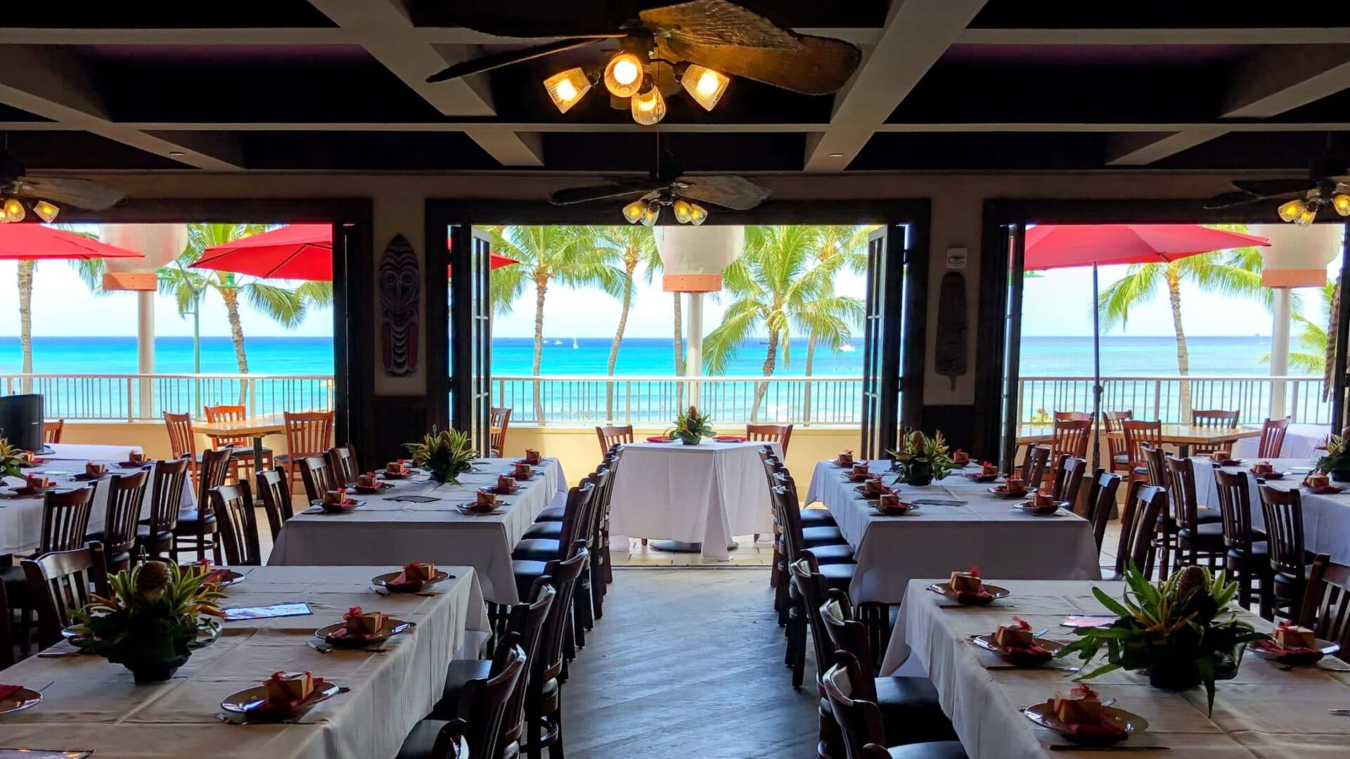 A large party table with white tablecloths and tropical flowers, set with an inside seating and an ocean view of the beach and coconut trees at Tiki's Grill & Bar.