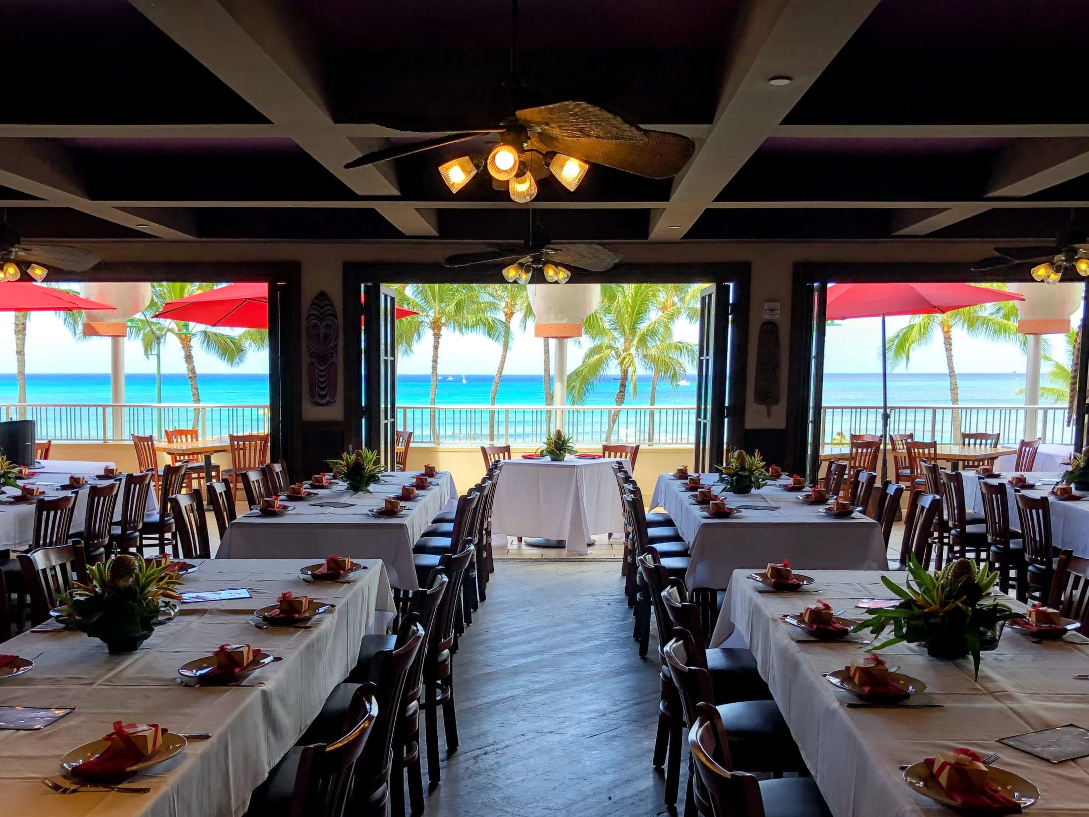 A large party table with white tablecloths and tropical flowers, set with an inside seating and an ocean view of the beach and coconut trees at Tiki's Grill & Bar.