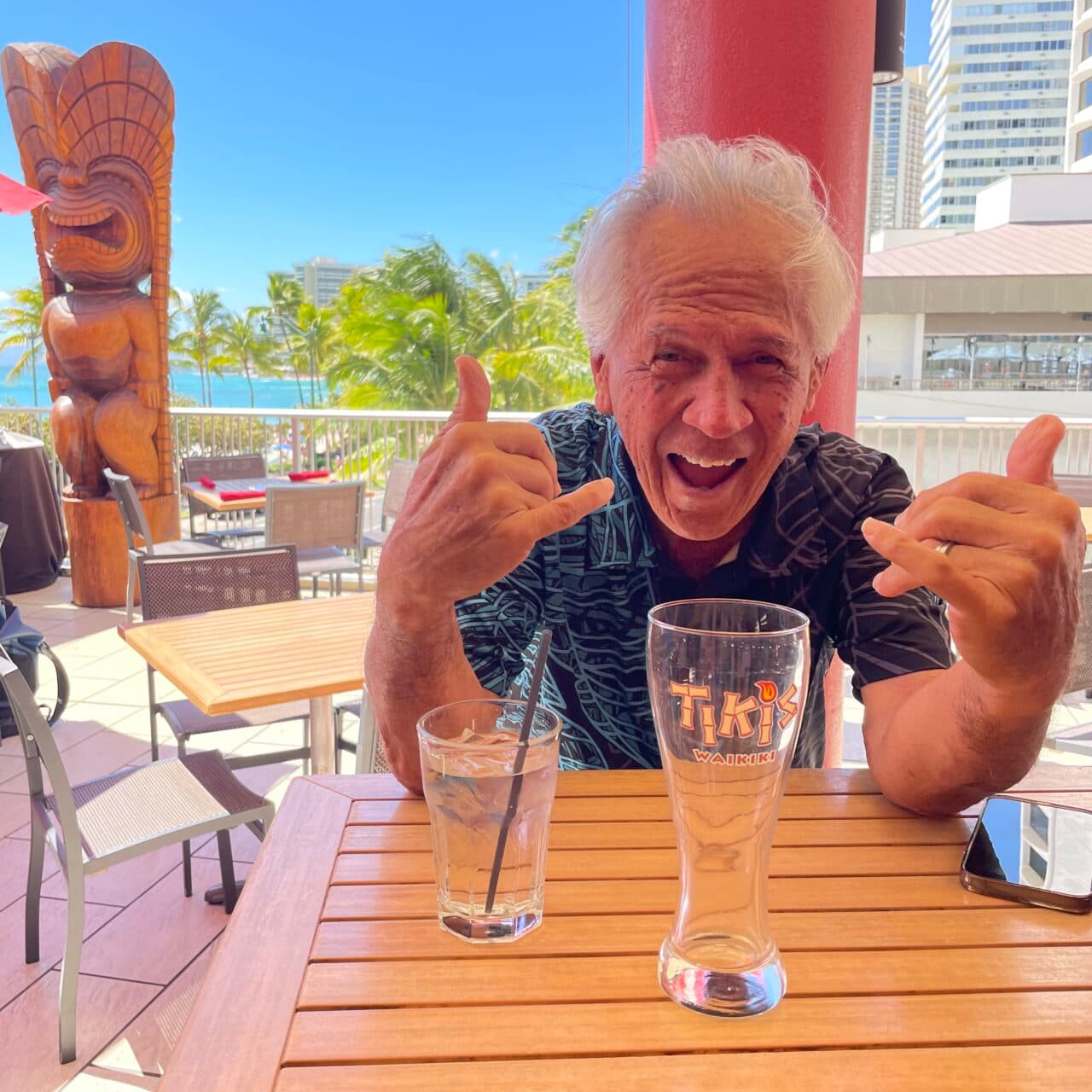 Kimo Kahoano flashes a shaka at Tiki’s Grill & Bar in Waikīkī with a large wooden tiki statue behind him.